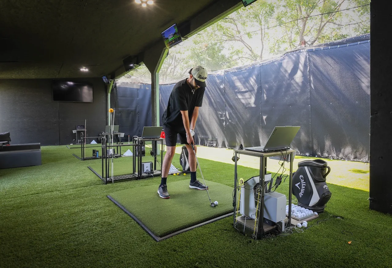Player hitting off turf in a covered outdoor bay with a laptop-connected launch monitor beside him