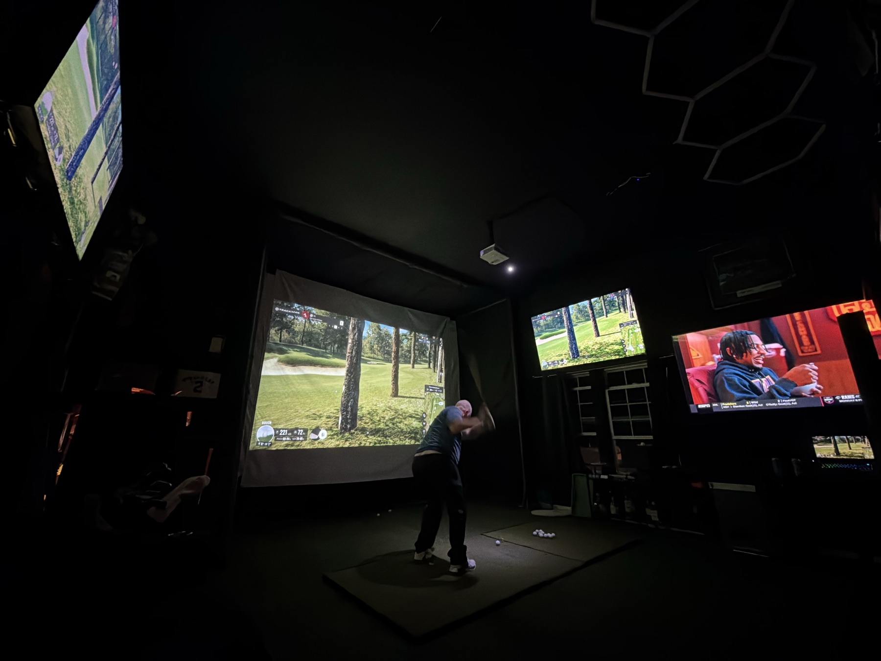 Golfer mid-swing in a blacked-out indoor simulator room with a projected course and TVs on the walls